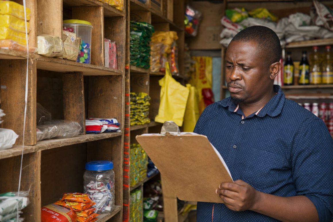 Shopkeeper confused at a clipboard next to empty shelves with mismatched stock counts